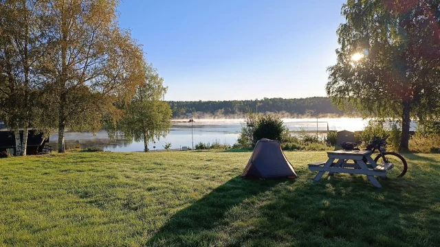 Zelt, Fahrrad und Picknicktisch auf einer Wiese am Seeufer bei Sonnenaufgang – Bikepacking Schweden am frühen Morgen.