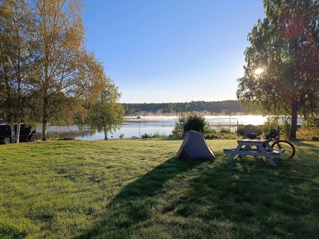 Zelt, Fahrrad und Picknicktisch auf einer Wiese am Seeufer bei Sonnenaufgang – Bikepacking Schweden am frühen Morgen.